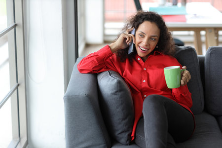 Smart and active latin woman sitting on sofa and using internet on computer with smart mobile phone while drinking coffee in living room. Lifestyle with technologyの写真素材