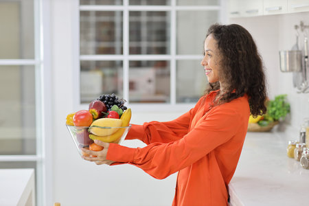 Happy latin woman standing and holding fruits basket while smiling and looking at camera in kitchen. Young female preparing delicious meal at home. Healthy and lifestyle conceptの写真素材