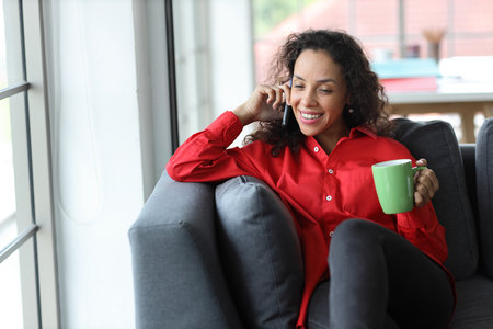 Smart and active latin woman sitting on sofa and using internet on computer with smart mobile phone while drinking coffee in living room. Lifestyle with technologyの写真素材