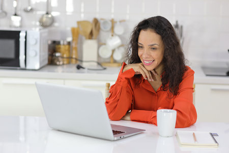 Smart and active latin woman sitting and using internet on computer while using smart mobile phone with a cup of coffee in kitchen. Anywhere and work. Lifestyle with technologyの写真素材