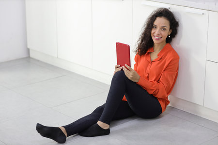 Happy latin woman sitting and using internet on tablet in kitchen room. Businesswoman looking at camera with smiling. Lifestyle with technologyの写真素材