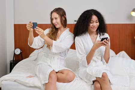 Young happy diversity ethnic beautiful woman in bathrobe sit on bed and playing social media on mobile smart phone and having fun together. Women lesbian couple daily LGBTQ lifestyle and love concept.の写真素材