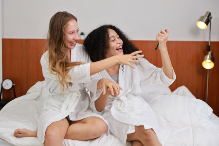 Young happy diversity ethnic beautiful woman in bathrobe sit on bed and playing social media on mobile smart phone and having fun together. Women lesbian couple daily LGBTQ lifestyle and love concept.の写真素材