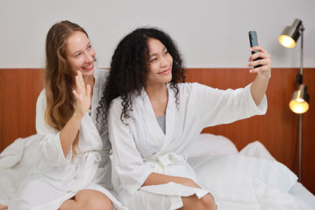 Young happy diversity ethnic beautiful woman in bathrobe sit on bed and playing social media on mobile smart phone and having fun together. Women lesbian couple daily LGBTQ lifestyle and love concept.の写真素材