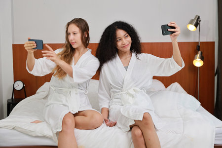 Young happy diversity ethnic beautiful woman in bathrobe sit on bed and playing social media on mobile smart phone and having fun together. Women lesbian couple daily LGBTQ lifestyle and love concept.の写真素材