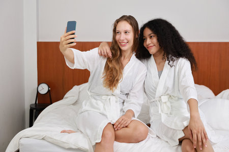 Young happy diversity ethnic beautiful woman in bathrobe sit on bed and playing social media on mobile smart phone and having fun together. Women lesbian couple daily LGBTQ lifestyle and love concept.の写真素材
