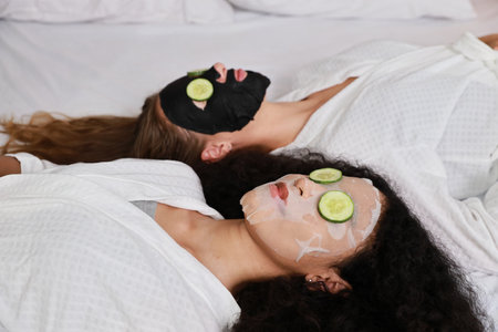 Young happy diversity ethnic beautiful woman in bathrobe lying on bed and doing cosmetic facial mask for skin care and treatment with cucumber slice. Women lesbian couple daily LGBTQ lifestyle conceptの写真素材