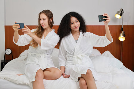 Young happy diversity ethnic beautiful woman in bathrobe sit on bed and playing social media on mobile smart phone and having fun together. Women lesbian couple daily LGBTQ lifestyle and love concept.の写真素材