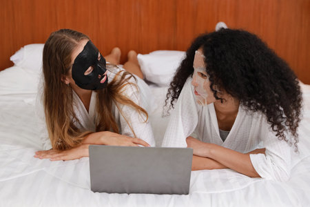 Young diversity ethnic beautiful woman in bathrobe playing social media on computer while doing cosmetic facial mask for skin care and treatment. Women lesbian couple daily LGBTQ lifestyle concept.の写真素材