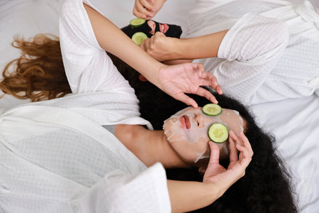 Young happy diversity ethnic beautiful woman in bathrobe lying on bed and doing cosmetic facial mask for skin care and treatment with cucumber slice. Women lesbian couple daily LGBTQ lifestyle conceptの写真素材