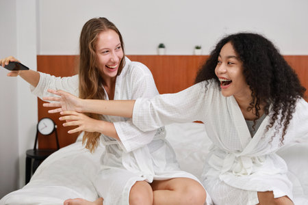 Young happy diversity ethnic beautiful woman in bathrobe sit on bed and playing social media on mobile smart phone and having fun together. Women lesbian couple daily LGBTQ lifestyle and love concept.の写真素材
