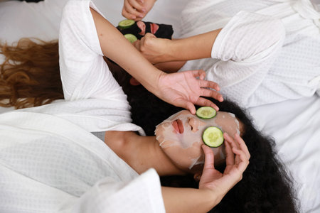 Young happy diversity ethnic beautiful woman in bathrobe lying on bed and doing cosmetic facial mask for skin care and treatment with cucumber slice. Women lesbian couple daily LGBTQ lifestyle conceptの写真素材