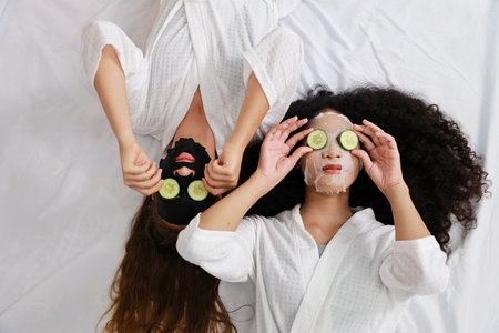 Young happy diversity ethnic beautiful woman in bathrobe lying on bed and doing cosmetic facial mask for skin care and treatment with cucumber slice. Women lesbian couple daily LGBTQ lifestyle conceptの写真素材