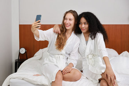 Young happy diversity ethnic beautiful woman in bathrobe sit on bed and playing social media on mobile smart phone and having fun together. Women lesbian couple daily LGBTQ lifestyle and love concept.の写真素材