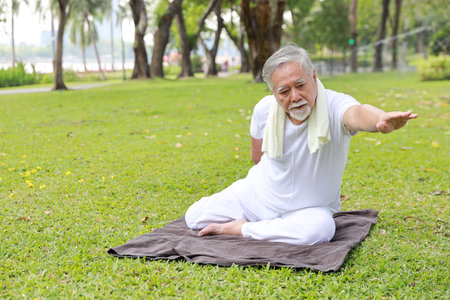 Asian senior man practice yoga exercise, stretching, tai chi tranining and meditation while sitting on yoga mat for healthy in park outdoor after retirement. Happy elderly outdoor lifestyle conceptの写真素材