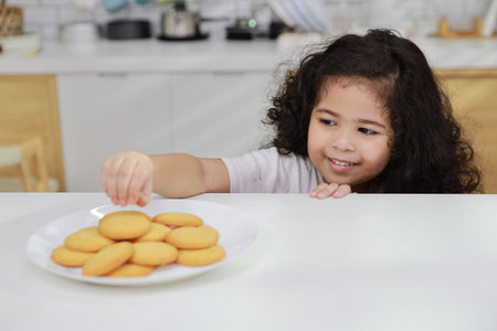 Young hispanic currly hair girl sitting on table and drinking milk while eating vanilla cookies for breakfast with enjoying time. Tasty food and delicious food with happy meal lifestyle kid conceptの写真素材