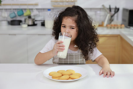 Young hispanic currly hair girl sitting on table and drinking milk while eating vanilla cookies for breakfast with enjoying time. Tasty food and delicious food with happy meal lifestyle kid conceptの写真素材