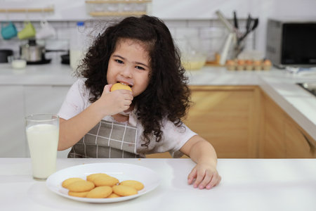 Young hispanic currly hair girl sitting on table and drinking milk while eating vanilla cookies for breakfast with enjoying time. Tasty food and delicious food with happy meal lifestyle kid conceptの写真素材