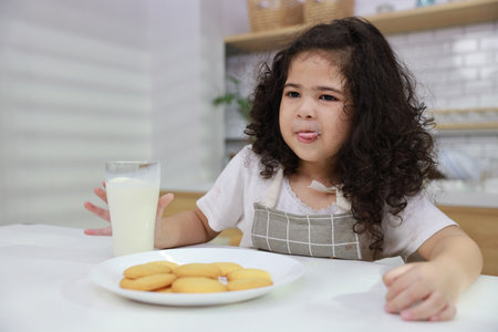 Young hispanic currly hair girl sitting on table and drinking milk while eating vanilla cookies for breakfast with enjoying time. Tasty food and delicious food with happy meal lifestyle kid conceptの写真素材