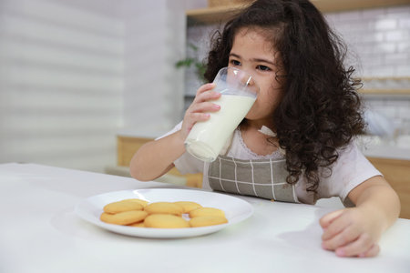 Kid hispanic currly hair girl sitting on table and drinking milk while eating vanilla cookies for breakfast with enjoying time. Tasty food and delicious food with happy meal lifestyle kid conceptの写真素材