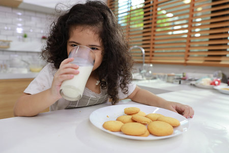 Kid hispanic currly hair girl sitting on table and drinking milk while eating vanilla cookies for breakfast with enjoying time. Tasty food and delicious food with happy meal lifestyle kid conceptの写真素材