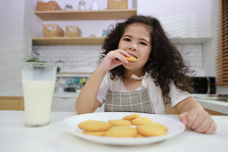 Kid hispanic currly hair girl sitting on table and drinking milk while eating vanilla cookies for breakfast with enjoying time. Tasty food and delicious food with happy meal lifestyle kid conceptの写真素材