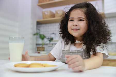 Young hispanic currly hair girl sitting on table and drinking milk while eating vanilla cookies for breakfast with enjoying time. Tasty food and delicious food with happy meal lifestyle kid conceptの写真素材