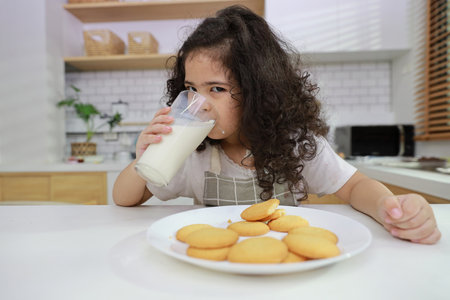 Young hispanic currly hair girl sitting on table and drinking milk while eating vanilla cookies for breakfast with enjoying time. Tasty food and delicious food with happy meal lifestyle kid conceptの写真素材