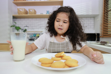 Young hispanic currly hair girl sitting on table and drinking milk while eating vanilla cookies for breakfast with enjoying time. Tasty food and delicious food with happy meal lifestyle kid conceptの写真素材