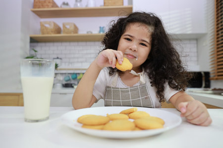 Young hispanic currly hair girl sitting on table and drinking milk while eating vanilla cookies for breakfast with enjoying time. Tasty food and delicious food with happy meal lifestyle kid conceptの写真素材