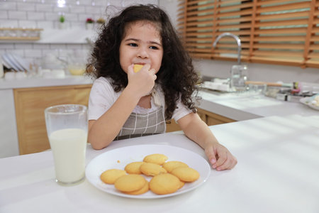 Kid hispanic currly hair girl sitting on table and drinking milk while eating vanilla cookies for breakfast with enjoying time. Tasty food and delicious food with happy meal lifestyle kid conceptの写真素材