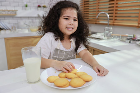 Young hispanic currly hair girl sitting on table and drinking milk while eating vanilla cookies for breakfast with enjoying time. Tasty food and delicious food with happy meal lifestyle kid conceptの写真素材