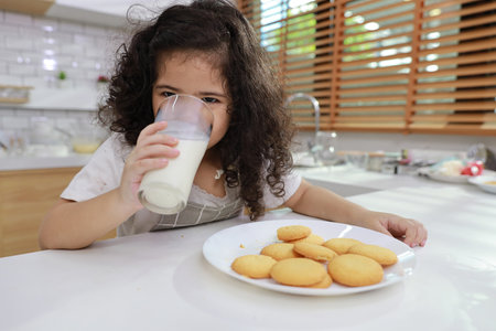 Young hispanic currly hair girl sitting on table and drinking milk while eating vanilla cookies for breakfast with enjoying time. Tasty food and delicious food with happy meal lifestyle kid conceptの写真素材
