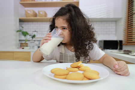 Kid hispanic currly hair girl sitting on table and drinking milk while eating vanilla cookies for breakfast with enjoying time. Tasty food and delicious food with happy meal lifestyle kid conceptの写真素材