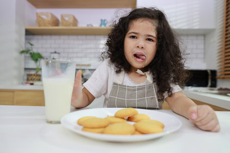 Young hispanic currly hair girl sitting on table and drinking milk while eating vanilla cookies for breakfast with enjoying time. Tasty food and delicious food with happy meal lifestyle kid conceptの写真素材