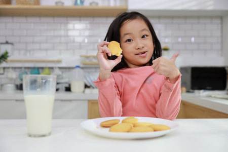 Kid asian black hair girl showing thumb up while drinking milk and eating vanilla cookies for breakfast with enjoying time. Tasty food and delicious food with happy meal lifestyle kid conceptの写真素材