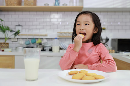 Kid asian black hair girl sitting on table and drinking milk while eating vanilla cookies for breakfast with enjoying time. Tasty food and delicious food with happy meal lifestyle kid conceptの写真素材