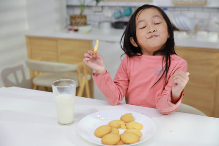 Kid asian black hair girl sitting on table and drinking milk while eating vanilla cookies for breakfast with enjoying time. Tasty food and delicious food with happy meal lifestyle kid conceptの写真素材