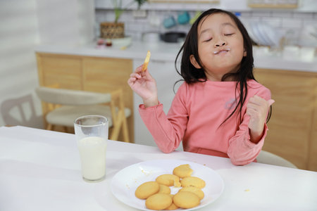 Kid asian black hair girl sitting on table and drinking milk while eating vanilla cookies for breakfast with enjoying time. Tasty food and delicious food with happy meal lifestyle kid conceptの写真素材