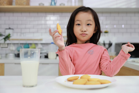 Kid asian black hair girl sitting on table and drinking milk while eating vanilla cookies for breakfast with enjoying time. Tasty food and delicious food with happy meal lifestyle kid conceptの写真素材