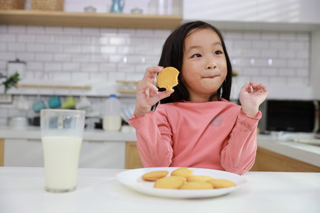 Kid asian black hair girl sitting on table and drinking milk while eating vanilla cookies for breakfast with enjoying time. Tasty food and delicious food with happy meal lifestyle kid conceptの写真素材