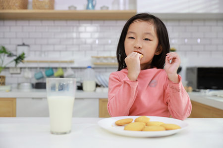 Kid asian black hair girl sitting on table and drinking milk while eating vanilla cookies for breakfast with enjoying time. Tasty food and delicious food with happy meal lifestyle kid conceptの写真素材