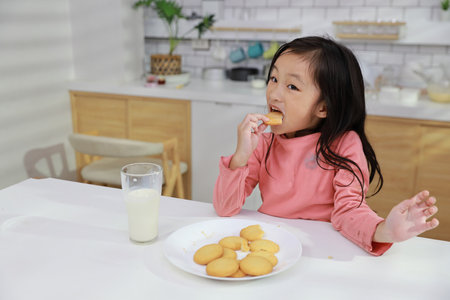 Kid asian black hair girl sitting on table and drinking milk while eating vanilla cookies for breakfast with enjoying time. Tasty food and delicious food with happy meal lifestyle kid conceptの写真素材