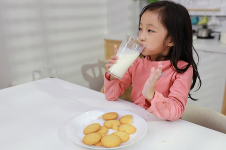 Kid asian black hair girl sitting on table and drinking milk while eating vanilla cookies for breakfast with enjoying time. Tasty food and delicious food with happy meal lifestyle kid conceptの写真素材