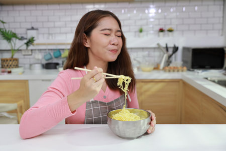 Portrait of young happy asian currly hair woman sitting on table and eating tasty and instant noodle for lunch in the kitchen with chopsticks and brick wall. Unhealthy and cheap food lifestyle conceptの写真素材