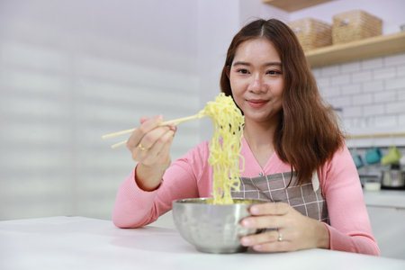 Portrait of young happy asian currly hair woman sitting on table and eating tasty and instant noodle for lunch in the kitchen with chopsticks and brick wall. Unhealthy and cheap food lifestyle conceptの写真素材