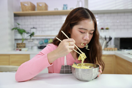 Portrait of young happy asian currly hair woman sitting on table and eating tasty and instant noodle for lunch in the kitchen with chopsticks and brick wall. Unhealthy and cheap food lifestyle conceptの写真素材