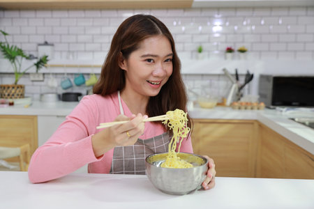 Portrait of young happy asian currly hair woman sitting on table and eating tasty and instant noodle for lunch in the kitchen with chopsticks and brick wall. Unhealthy and cheap food lifestyle conceptの写真素材