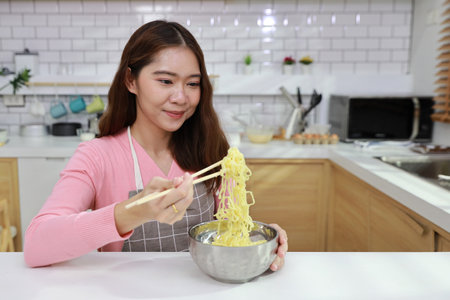 Portrait of young happy asian currly hair woman sitting on table and eating tasty and instant noodle for lunch in the kitchen with chopsticks and brick wall. Unhealthy and cheap food lifestyle conceptの写真素材