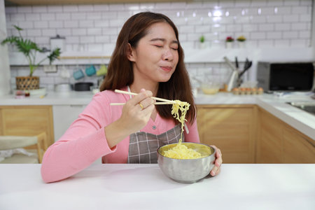 Portrait of young happy asian currly hair woman sitting on table and eating tasty and instant noodle for lunch in the kitchen with chopsticks and brick wall. Unhealthy and cheap food lifestyle conceptの写真素材